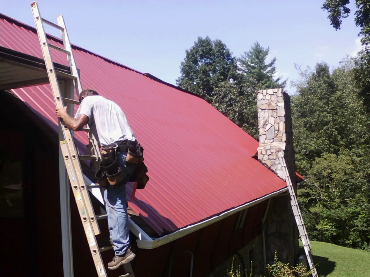 Roofer on a ladder beside a red metal roof and stone chimney — Soffit & Fascia Repair by Roofing Brothers in Old Hickory